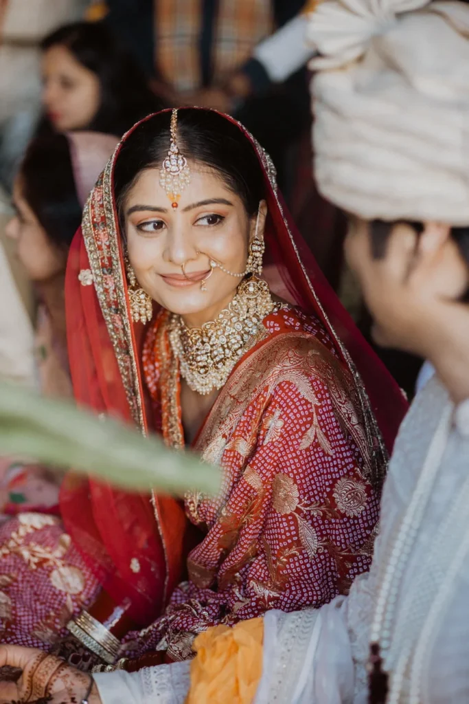 Bride smiling during the wedding ceremony while dressed in a traditional red lehenga with classic bridal jewelry and veil.