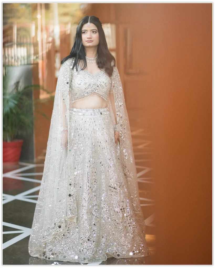 Bride wearing a shimmering silver lehenga with elegant jewelry, standing gracefully in natural light.
