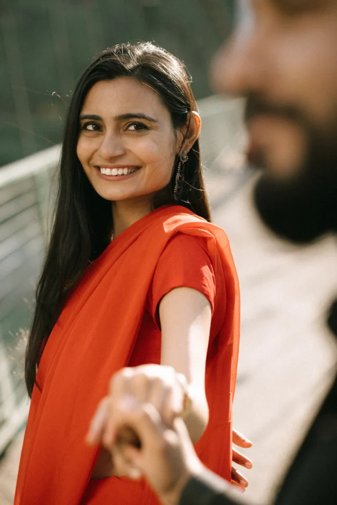 Smiling bride in a flowing red dress holding her partner's hand with soft natural light and shallow depth of field