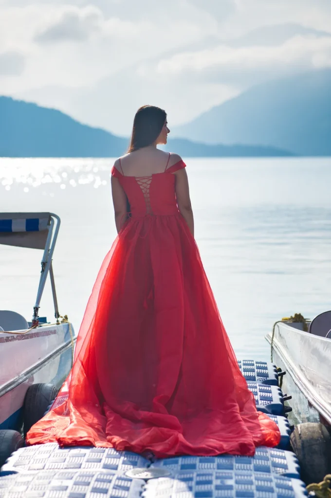 Woman in a flowing red gown standing by the water with scenic hills behind her.