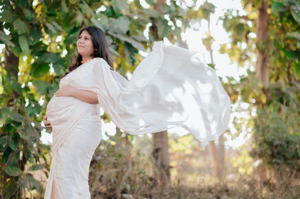 Graceful mother-to-be surrounded by natural greenery with a flowing saree creating a dreamy outdoor portrait