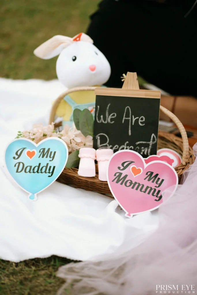 Pregnancy announcement setup with baby shoes, heart-shaped signs, and nursery props arranged for an outdoor photoshoot.