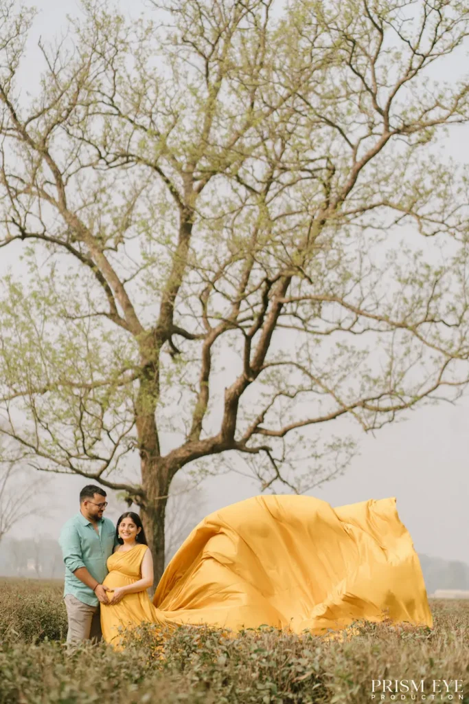 Expecting couple posing beneath a large tree with elegant flowing fabric creating a dramatic outdoor portrait.