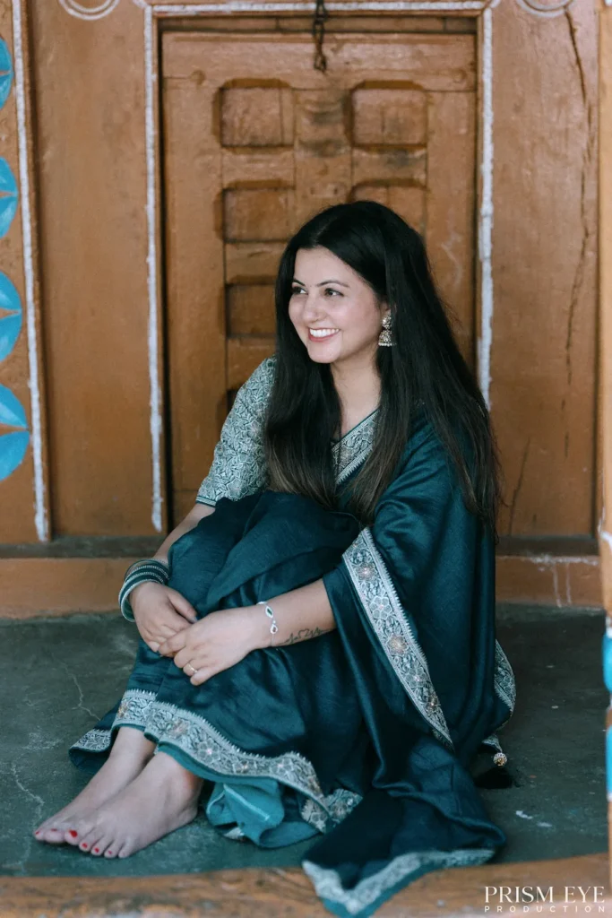 Smiling woman sitting in front of a heritage wooden doorway wearing a traditional green saree