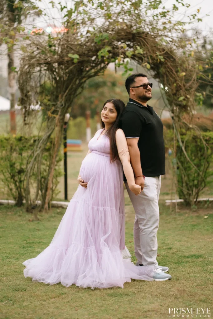 Expecting couple holding hands beneath a natural garden arch in an outdoor portrait.