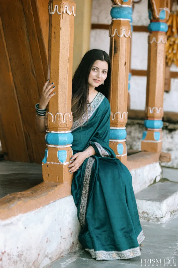 Woman in a green saree posing beside beautifully carved wooden pillars of a heritage village home