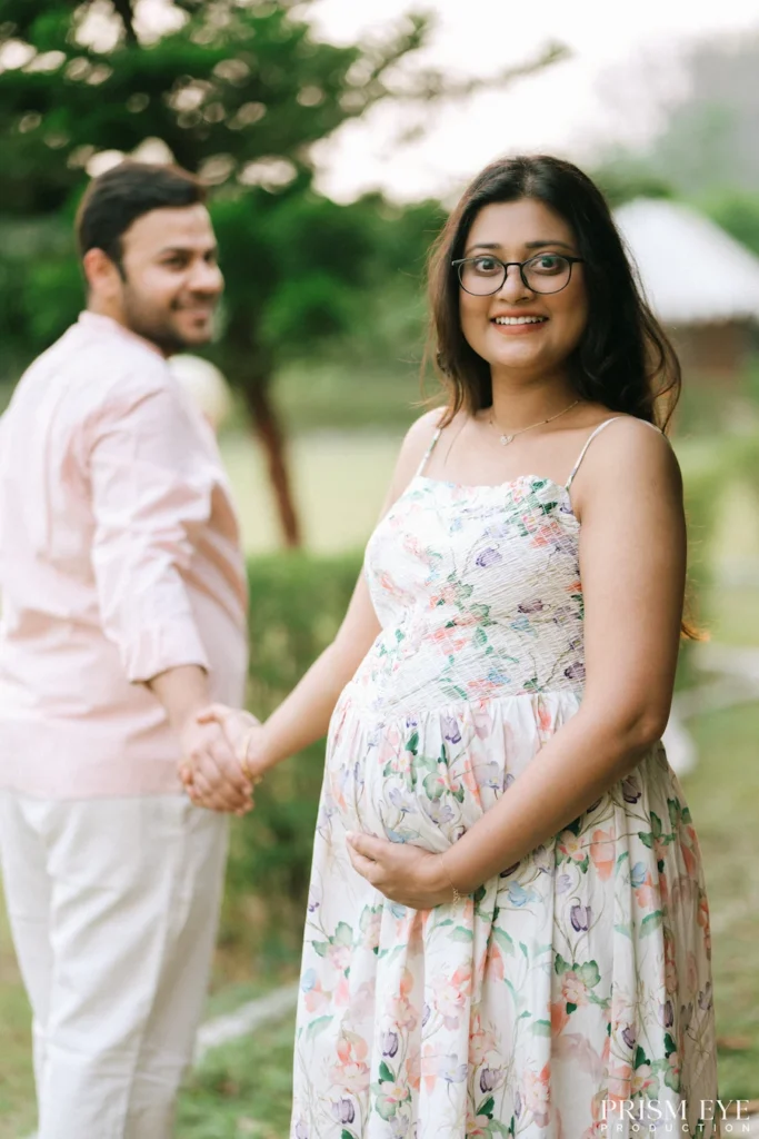 Smiling expectant mother gently holding her baby bump while her partner walks beside her outdoors.