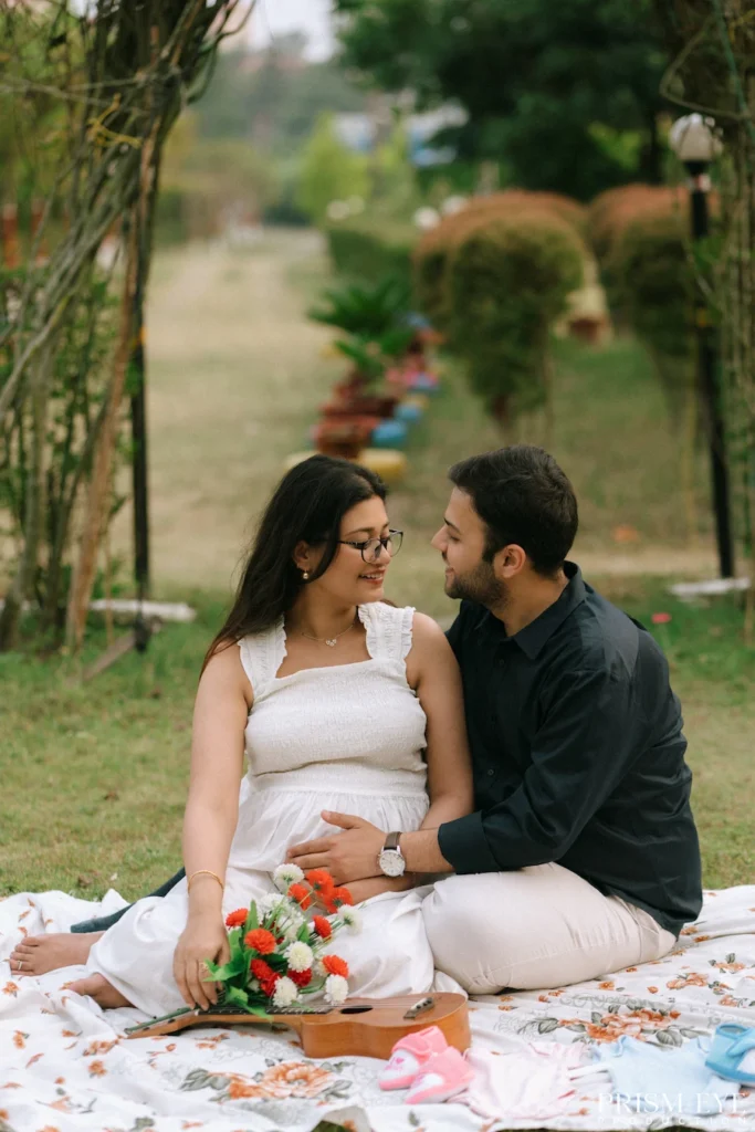 Expecting couple sitting together on a picnic blanket surrounded by baby essentials and flowers.