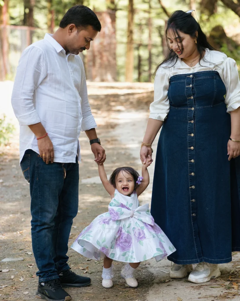 Parents helping their daughter take her first steps during a family photoshoot in a park.