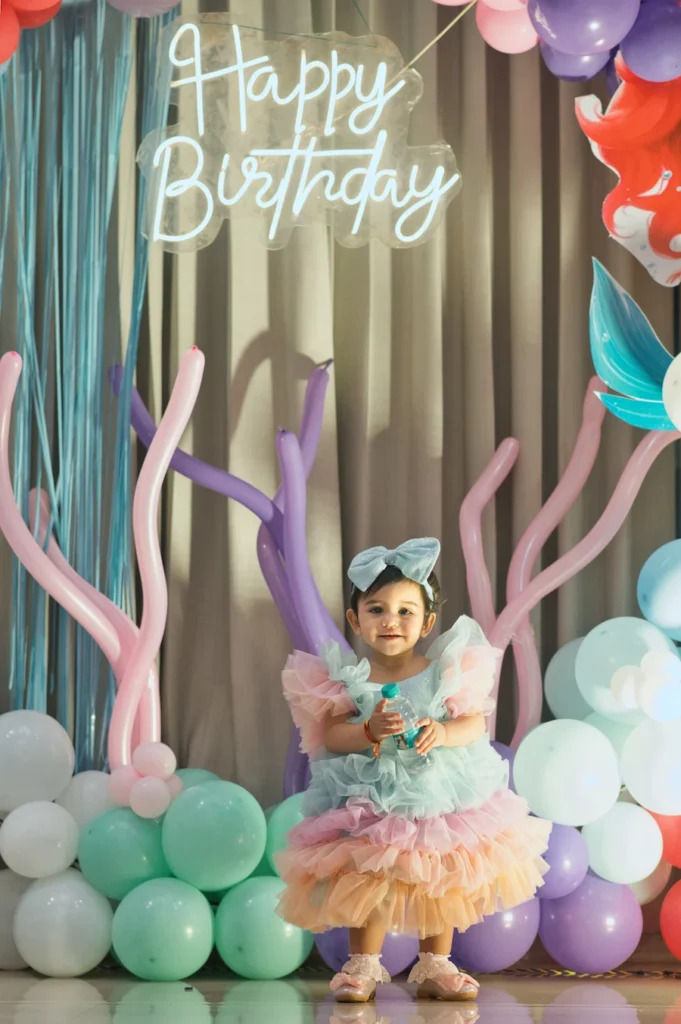 Toddler standing beneath a glowing Happy Birthday sign with pastel balloons.