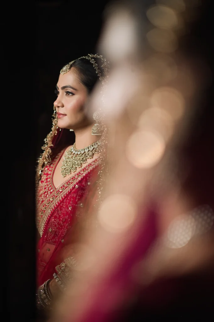 Bride posing in profile with soft foreground bokeh, highlighting elegant bridal jewelry and traditional red wedding attire.