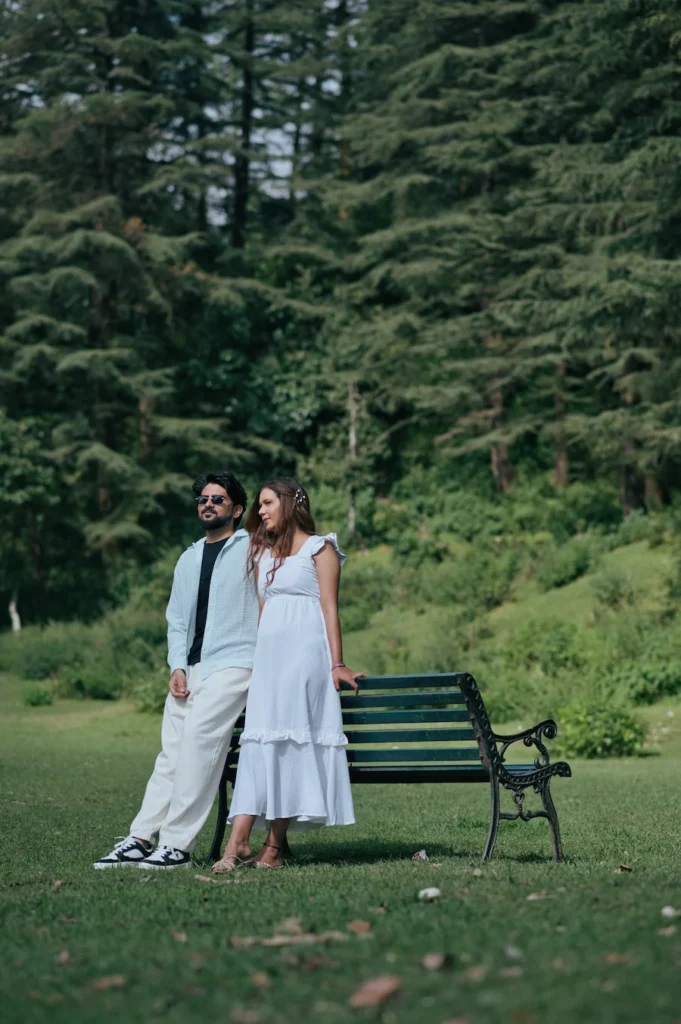 Couple standing beside a park bench surrounded by lush green trees.