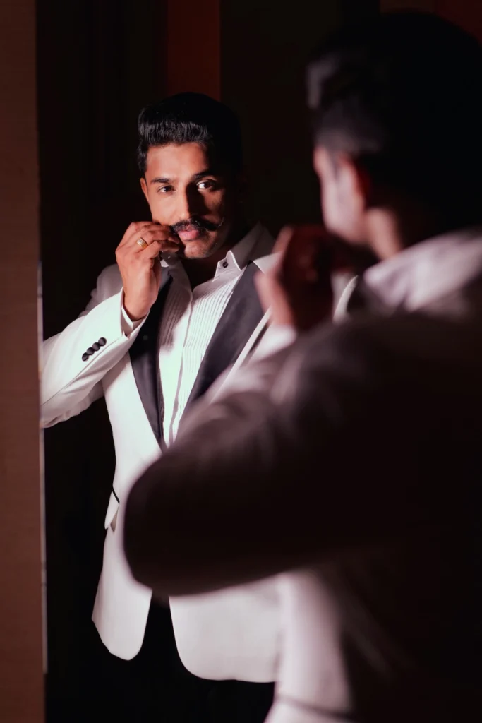 Groom in a white tuxedo jacket adjusting his mustache while looking at his reflection in a mirror.
