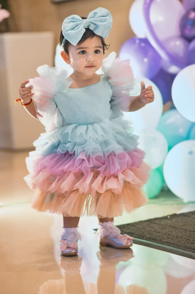 Alt Text: First birthday photoshoot of a baby girl in a colorful rainbow dress standing beside pastel balloons