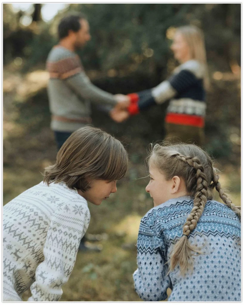 Two siblings sharing a playful moment while their parents hold hands in the background among the pine trees.