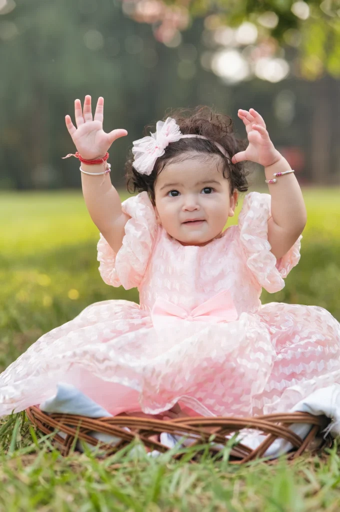 Baby girl in a pink birthday dress sitting in a wicker basket with hands raised during an outdoor portrait session
