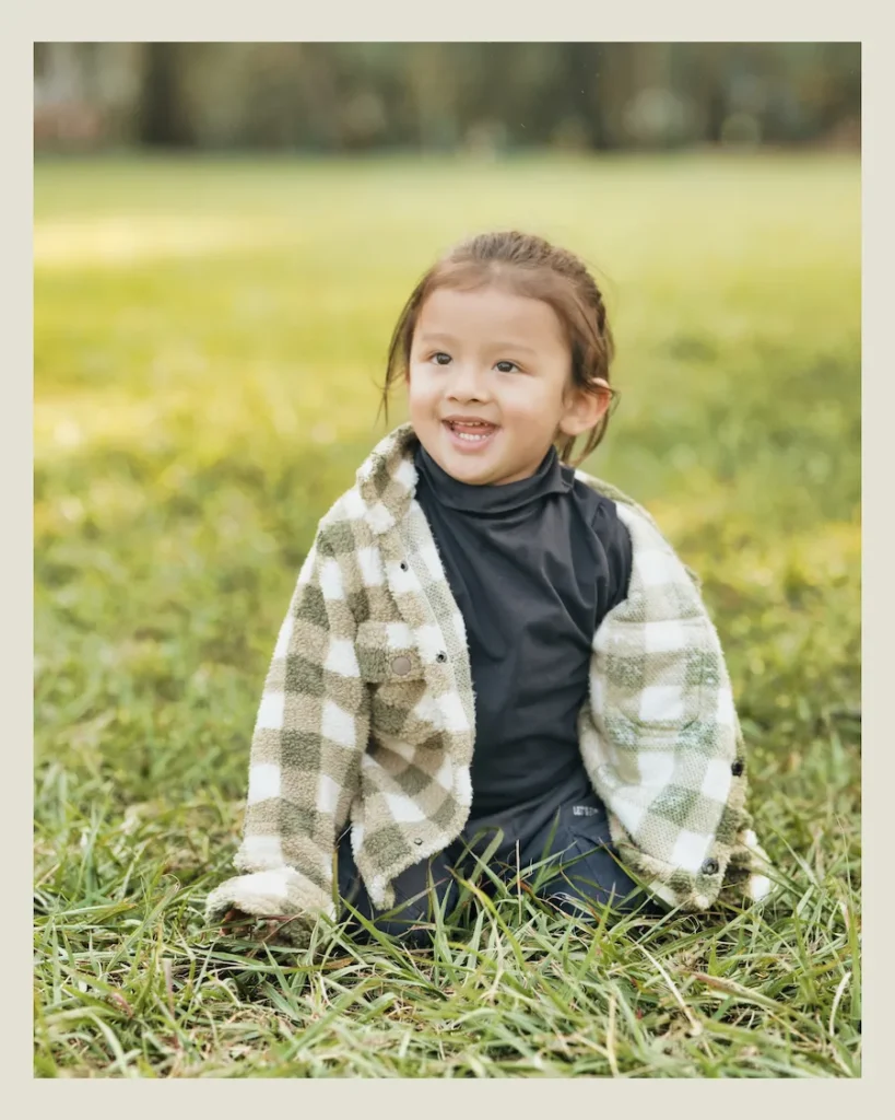 Smiling toddler sitting on green grass wearing a stylish jacket during a natural outdoor portrait