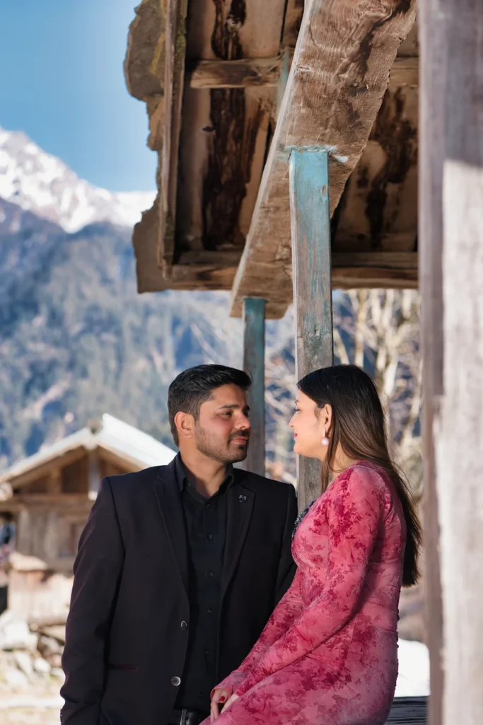 Couple portrait near a traditional mountain village house with snow-capped Himalayan peaks in the background.