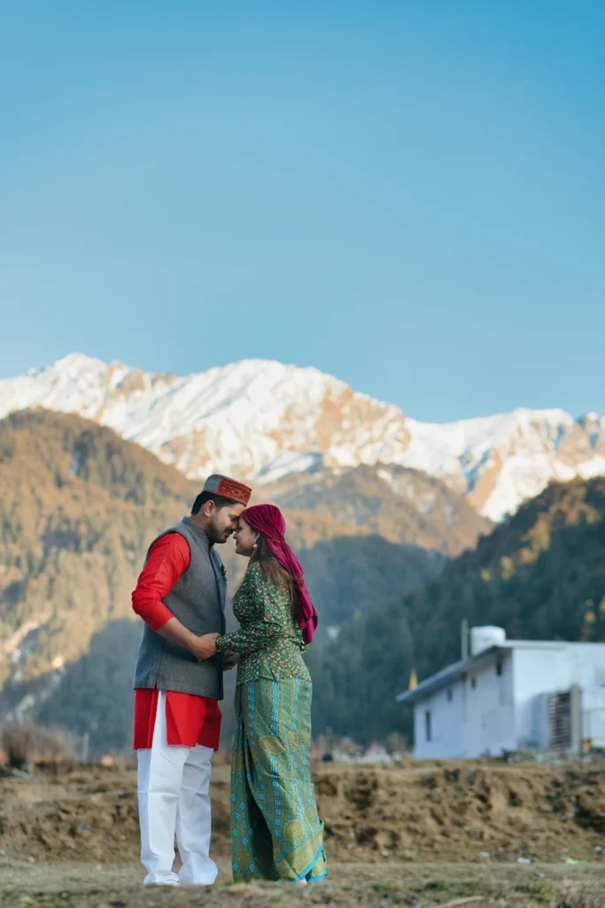 Couple dressed in traditional Pahadi attire posing against snow-capped Himalayan mountains during a cultural pre wedding session.