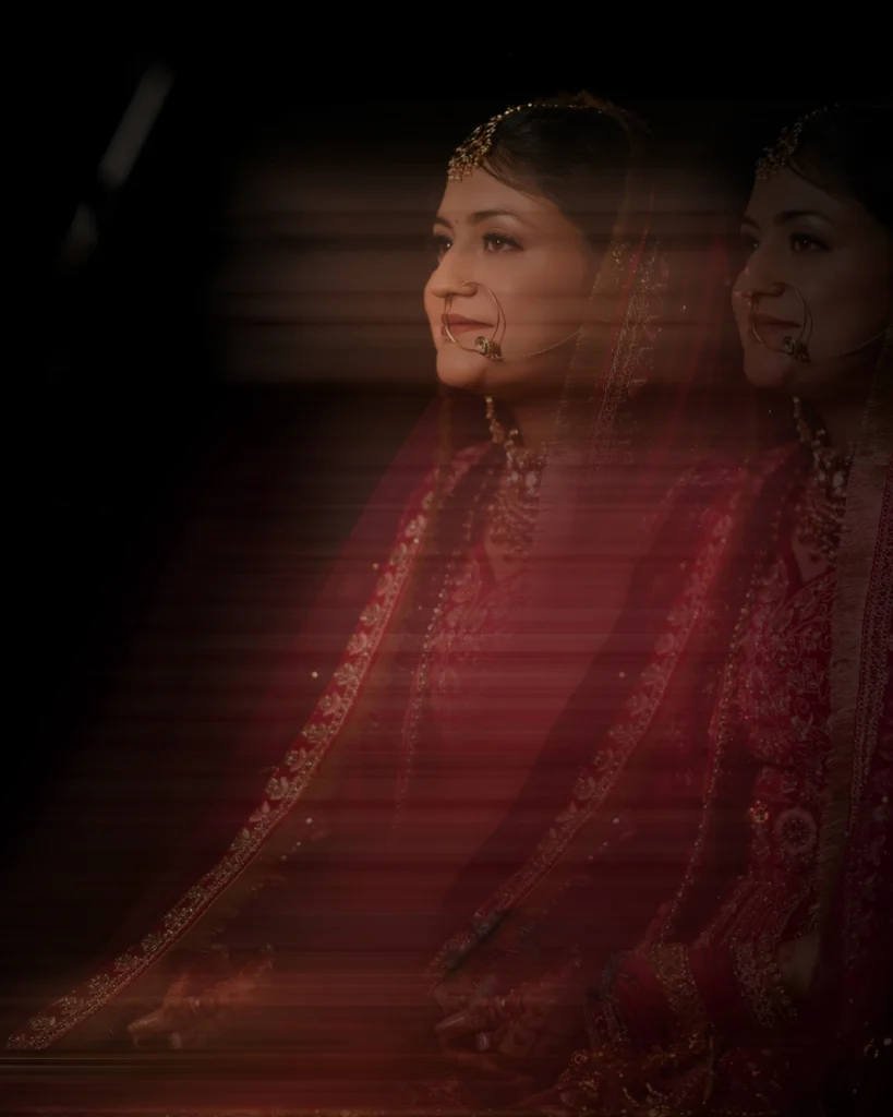 A vertical three-quarter creative bridal portrait featuring a double exposure and horizontal motion blur effect, showing a profile view of an Indian bride looking to the left with a smile, dressed in a red embroidered lehenga, a sheer veil, and traditional wedding jewelry against a dramatic dark background.
