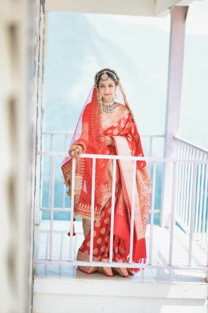 A graceful portrait of a bride dressed in a traditional red lehenga, standing on a balcony with a scenic mountain backdrop during her destination wedding in Uttarakhand, captured by Prism Eye Productions.