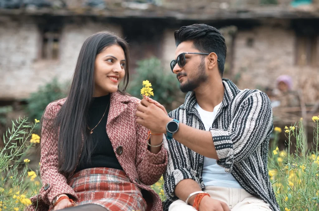 A playful and candid pre-wedding portrait of a couple sharing a lighthearted moment with a yellow flower in a rustic field, captured by Prism Eye Productions.