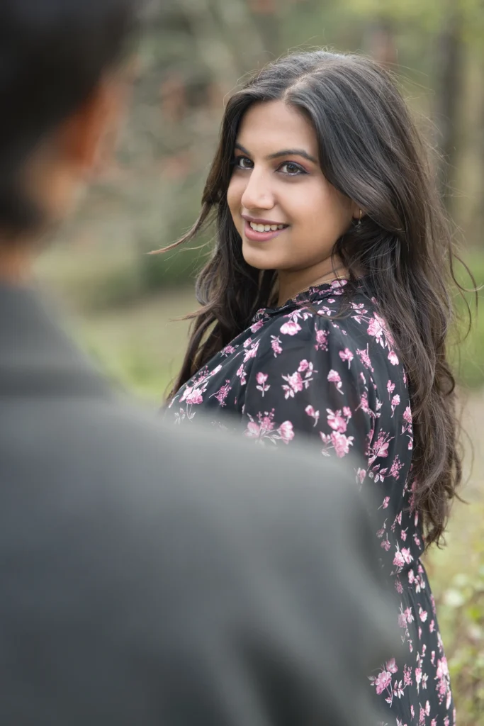 An over-the-shoulder shot of a man looking towards a smiling woman with long dark hair who looks back over her shoulder, wearing a black dress with a pink floral pattern in an outdoor wooded area.
