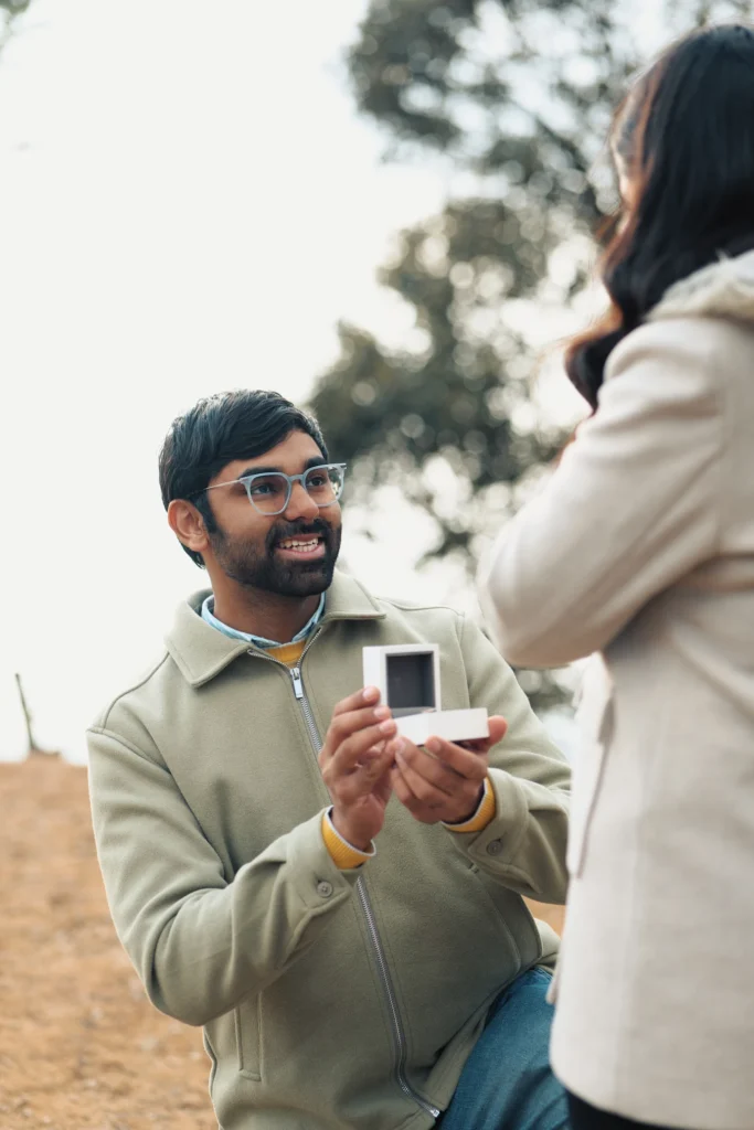 An over-the-shoulder shot of a young man with glasses down on one knee presenting a white engagement ring box to a woman in a light grey winter coat during an outdoor marriage proposal.