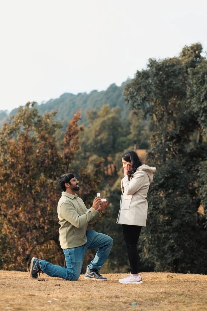 A young man down on one knee presents an engagement ring box to a woman who covers her mouth in surprise, set in an open grassy field with a backdrop of green forest trees.