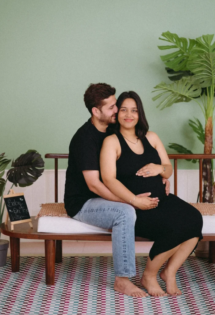 An expecting couple sitting on a wooden bench in a cozy indoor setting, with a small "Baby on the Way" chalkboard sign nearby.