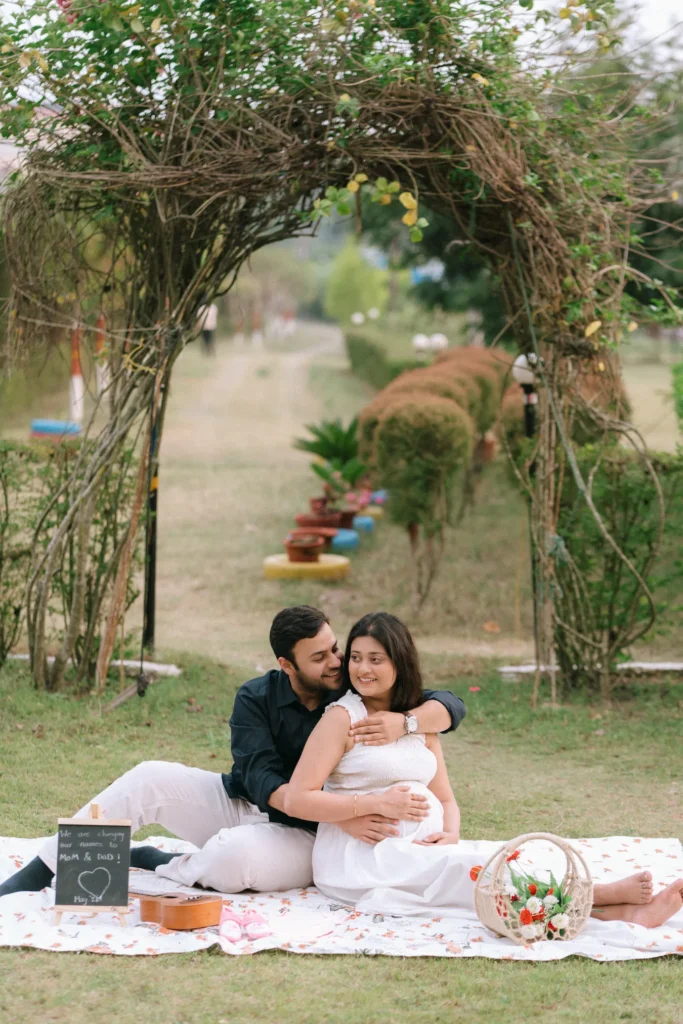 An expecting couple enjoying a romantic picnic in a garden, sitting on a floral blanket under a decorative archway.