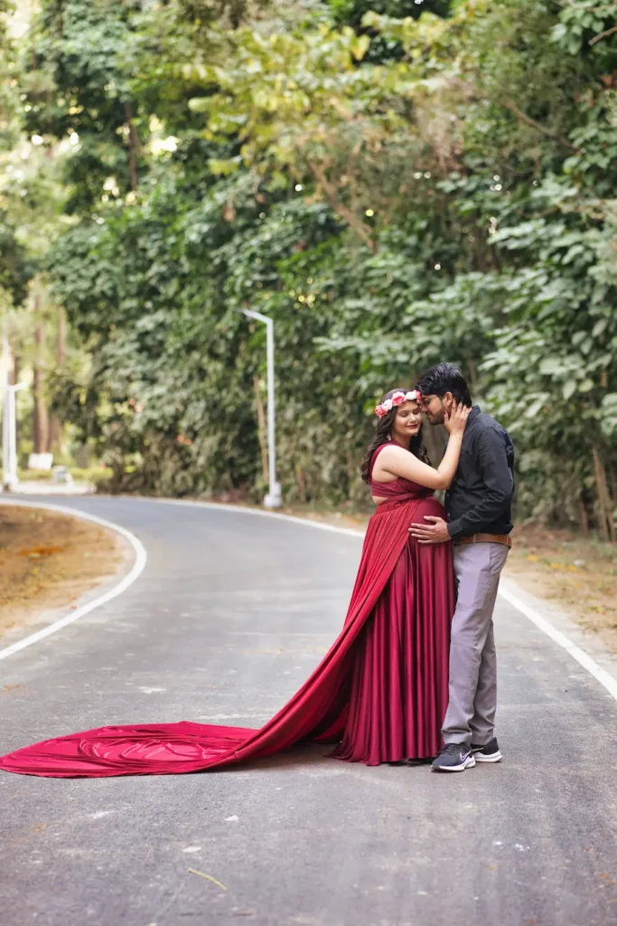 An expecting couple in a romantic embrace on a scenic forest road, with the mother wearing a flowing red maternity gown.