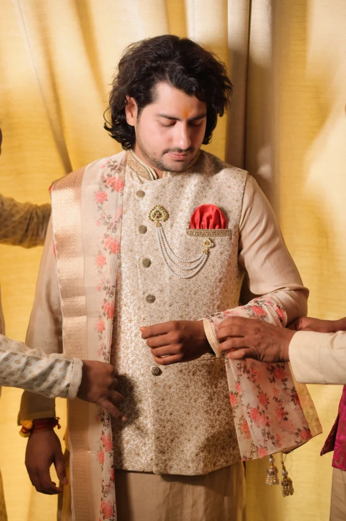 A close-up candid shot of a groom looking down while his traditional floral shawl is being adjusted by someone during his wedding preparation, set against a gold curtain backdrop.