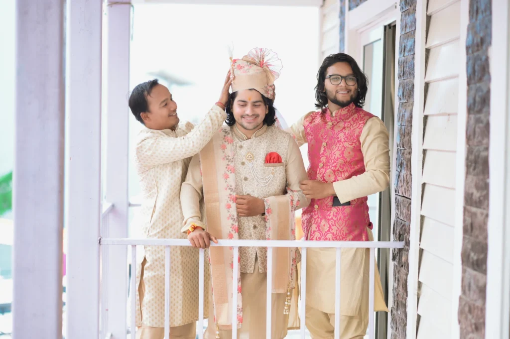 A groom in a traditional cream sherwani stands on a balcony while a friend adjusts his turban and another friend in a pink vest stands next to them smiling.