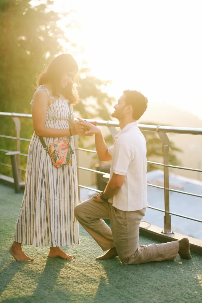 A young man down on one knee holds a woman's hand to place a ring on her finger on an outdoor terrace at sunset, with the woman standing barefoot in a striped sundress.