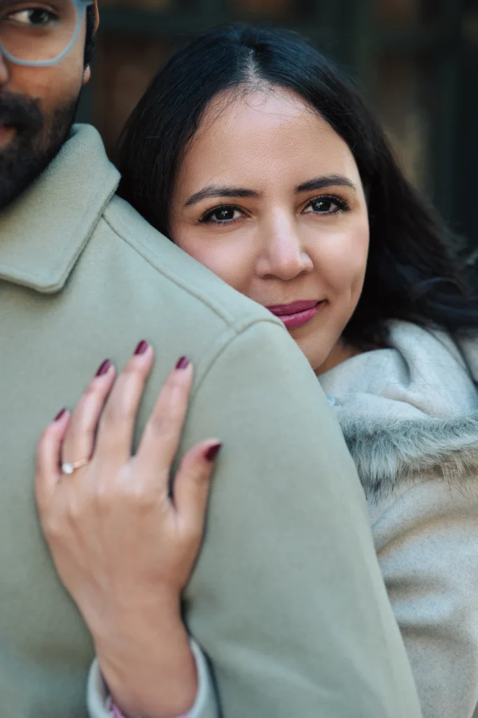 An extreme close-up portrait of a woman resting her face against a man's shoulder in a warm embrace, wearing a light grey coat with a fur collar while her hand shows an engagement ring on his sage green jacket.