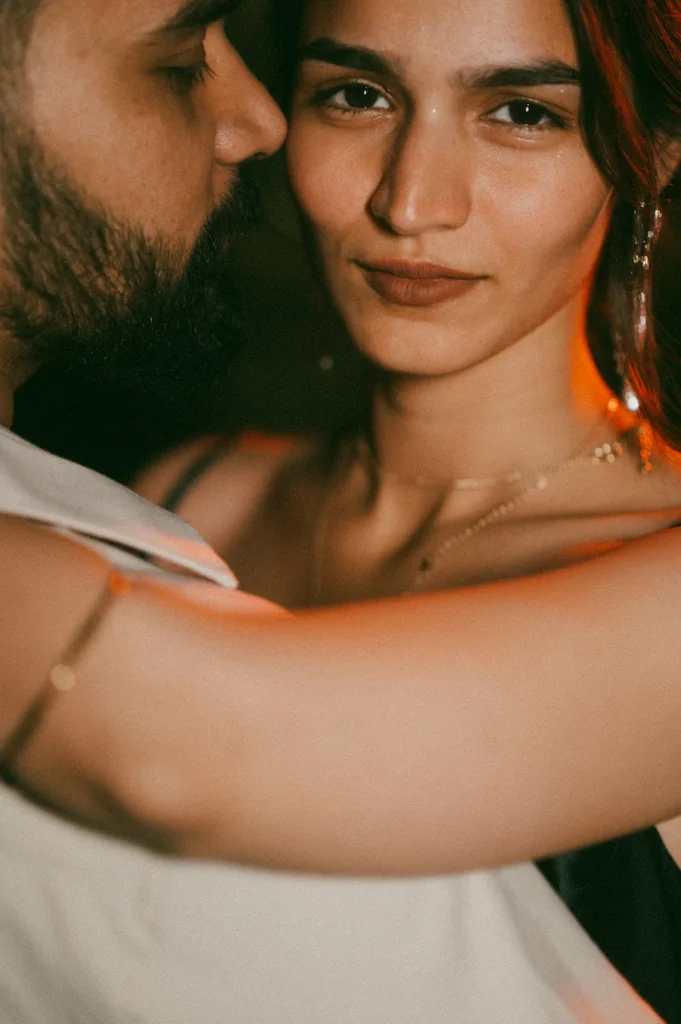A dramatic close-up vertical portrait of a woman looking intently at the camera while embracing a bearded man whose profile is close to her face, illuminated by a warm orange rim light at night.
