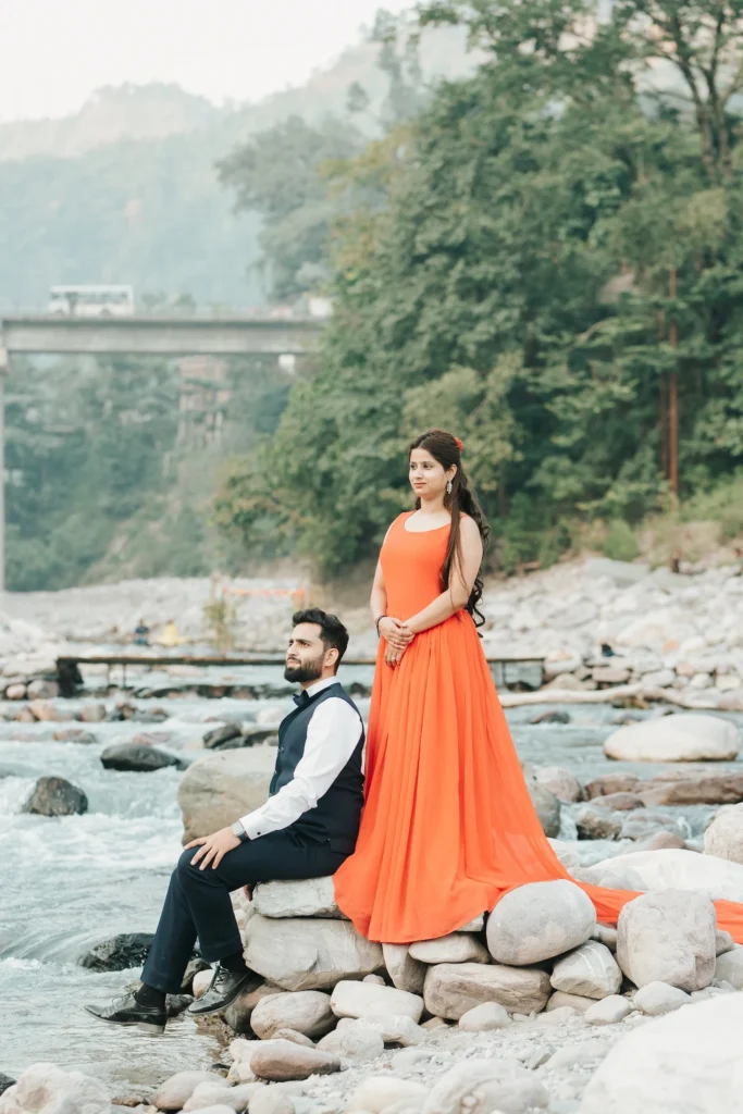 A couple posing together on the rocky riverbank in Rishikesh with a scenic bridge in the background.