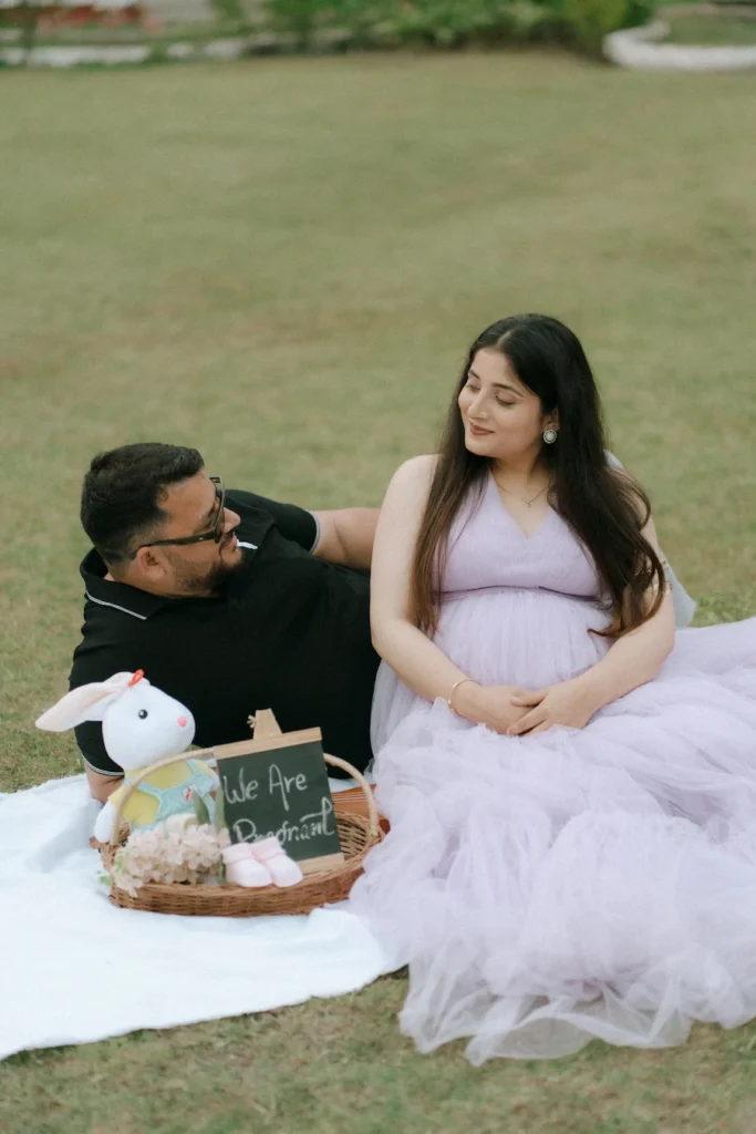 An expectant couple enjoying a garden picnic in Rishikesh, with the mother in a beautiful lavender dress and a "We Are Pregnant" sign nearby.