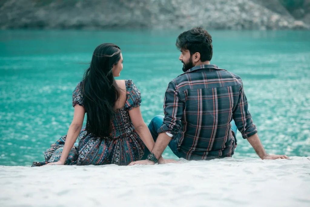 A couple sits side-by-side from a back angle on a white sandy beach, holding hands and looking at each other against the backdrop of vibrant turquoise river water.