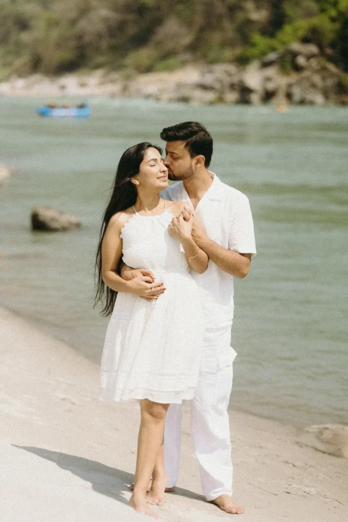 A young couple dressed in matching white outfits stands embracing on a sandy riverbank with the turquoise waters of the Ganges River flowing behind them during a photoshoot.