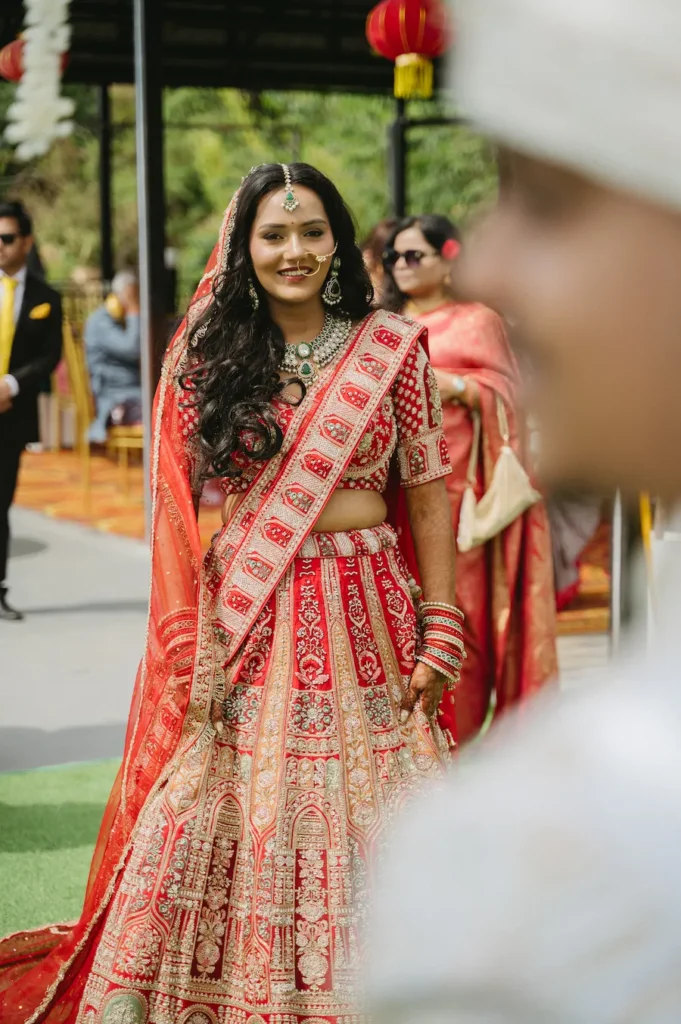 Smiling Indian bride in a red lehenga looking affectionately at her out-of-focus groom during their outdoor wedding ceremony.