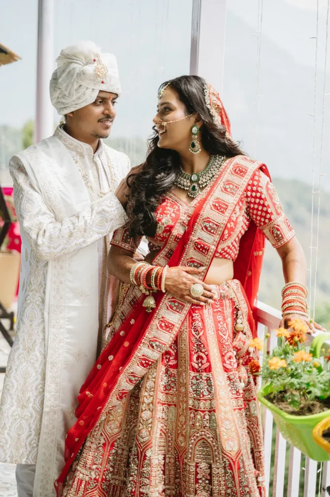 Smiling bride in a red lehenga and groom in a white sherwani sharing a candid moment on a balcony near Triyuginarayan Temple.
