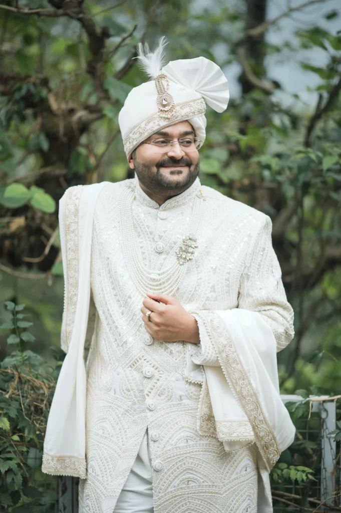 Groom in a traditional white sherwani and turban posing outdoors surrounded by greenery.