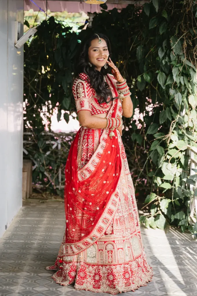 Smiling Indian bride in a red lehenga standing next to a lush green wall before her wedding at Triyuginarayan Temple.