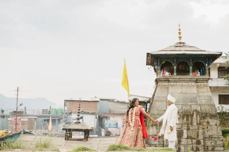 Bride and groom holding hands in traditional Indian wedding attire near an ancient stone shrine at Triyuginarayan Temple.
