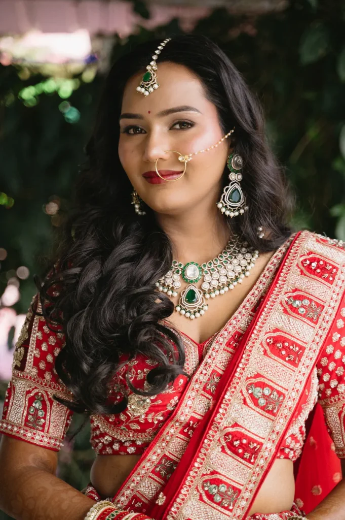 Close-up portrait of a smiling Indian bride in a red lehenga with elegant emerald and pearl jewelry near Triyuginarayan Temple.