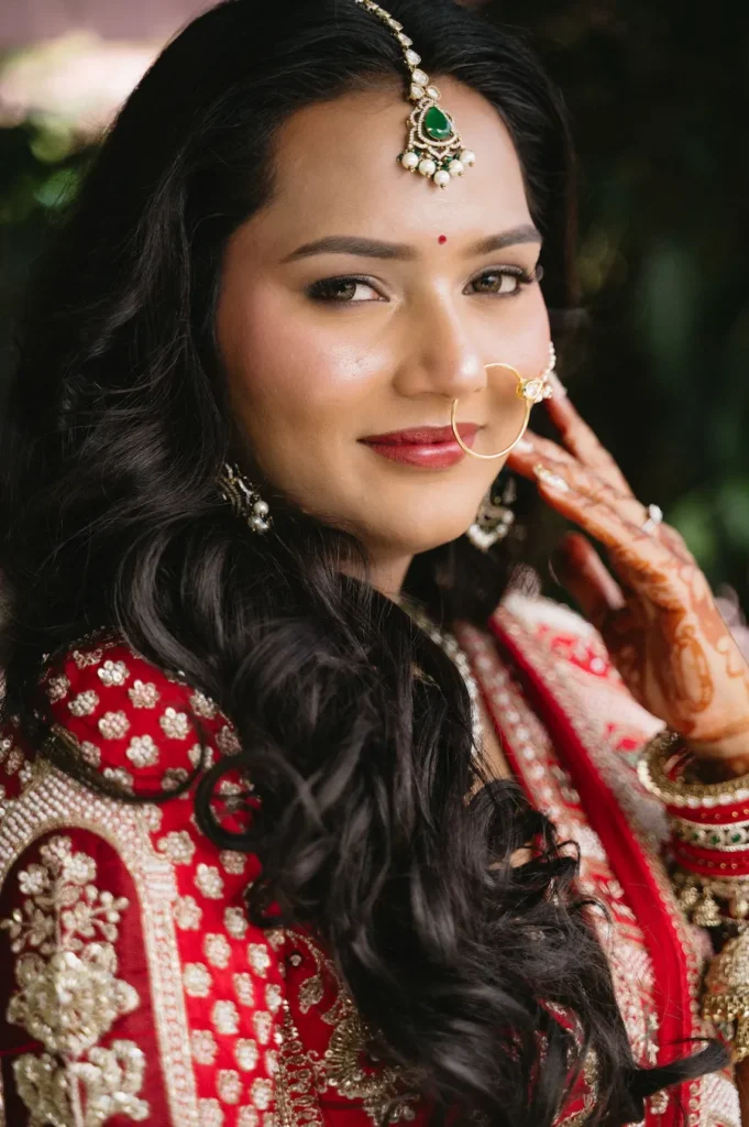 Smiling Indian bride with flowing dark hair, red lehenga, henna, and bridal jewelry posing near Triyuginarayan Temple.