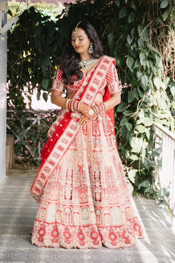Full-length portrait of a smiling Indian bride in a beautifully embroidered red lehenga standing against a green foliage backdrop near Triyuginarayan Temple.