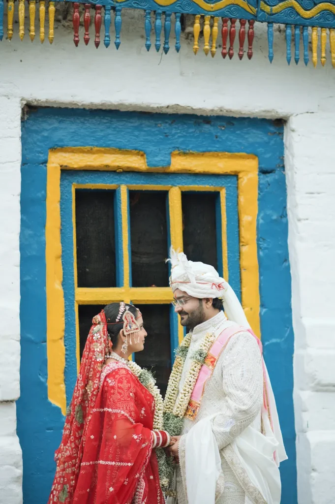 Bride and groom in traditional wedding attire standing together outside a colorful traditional house.
